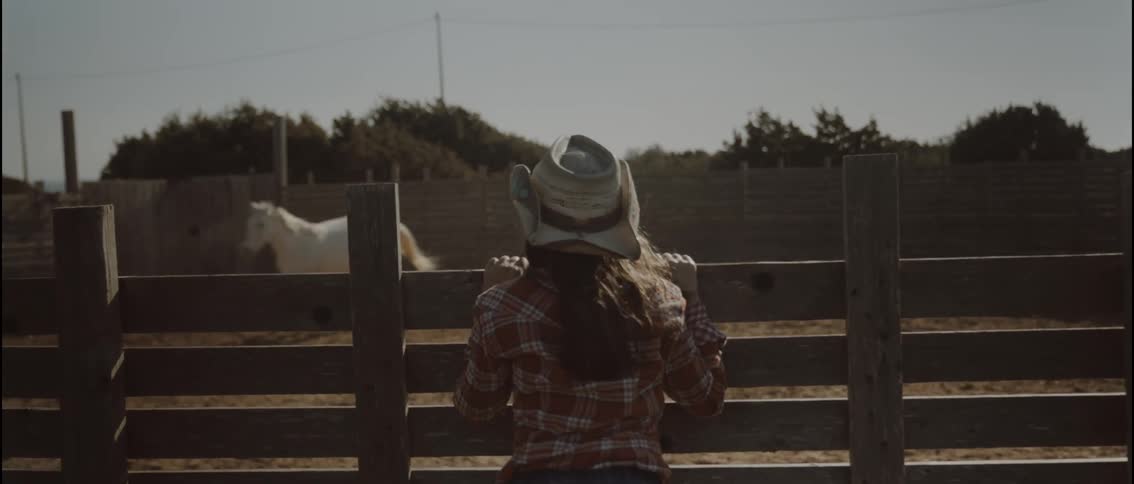 A girl with cowboy hat climbing a fence to watch a women chasing a horse around a pen