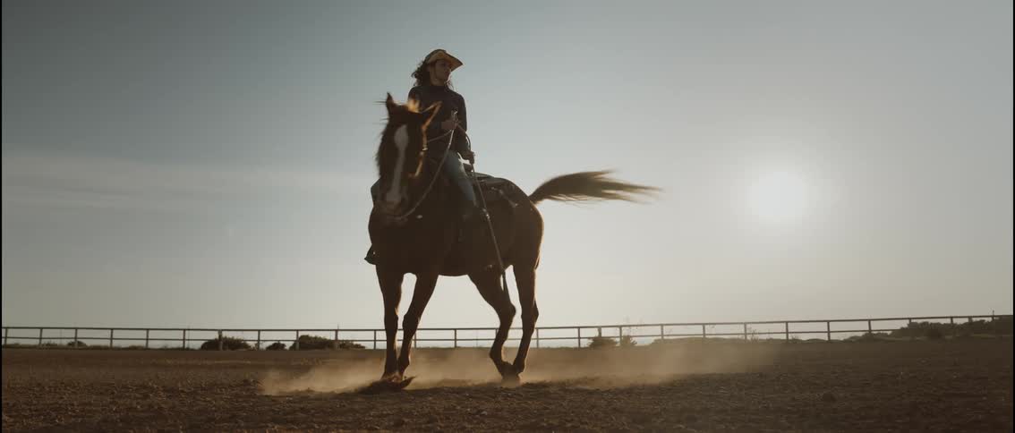 A young woman rides a horse at sunset wearing a cowboy hat as she turns in a full circle