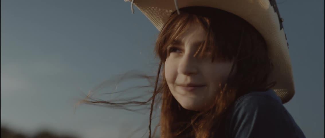 A young girl with long brown hair wearing a white cowboy hat and a denim shirt smiles while looking into the distance against a clear sky