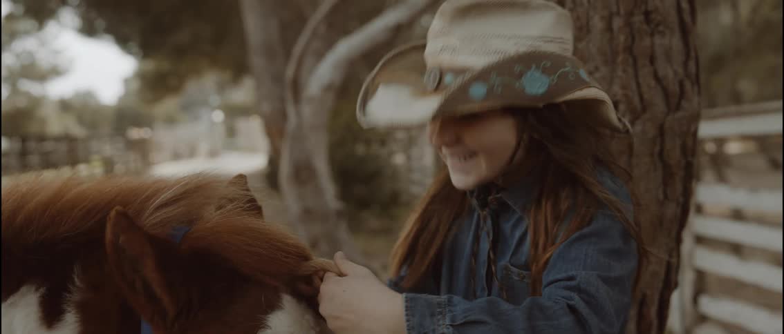 The video shows a young girl wearing a cowboy hat who is smiling while she braids the mane of a small horse the scene takes place outdoors in a ranchlike setting with trees and wooden fences visible in the softfocus background