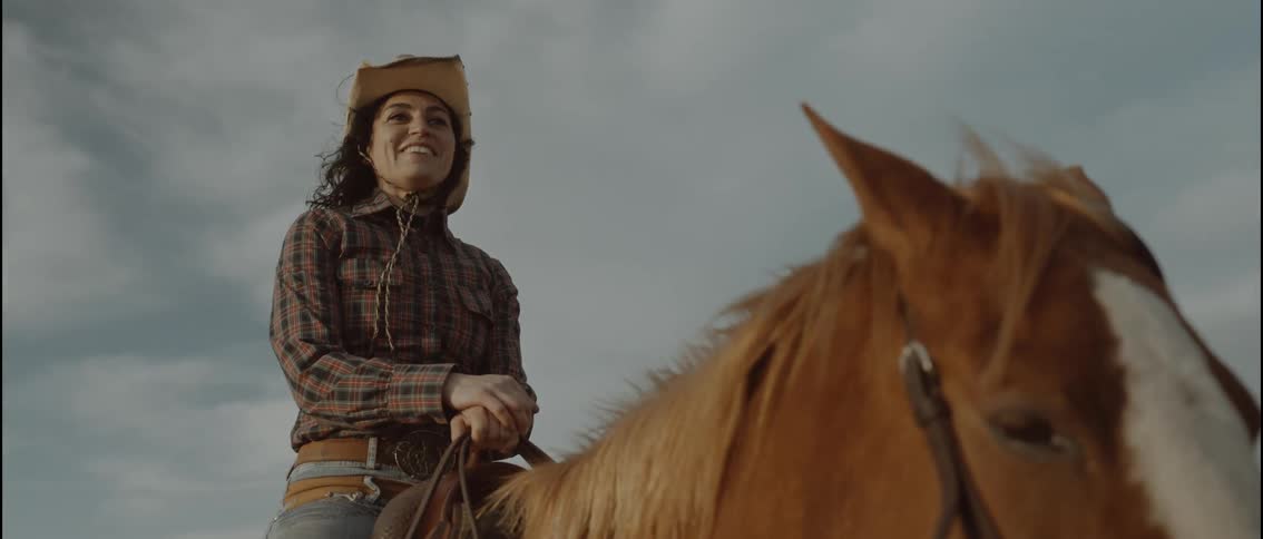 A smiling woman wearing a western hat and plaid shirt sits on a brown horse while looking toward the horizon against a cloudy sky during the day