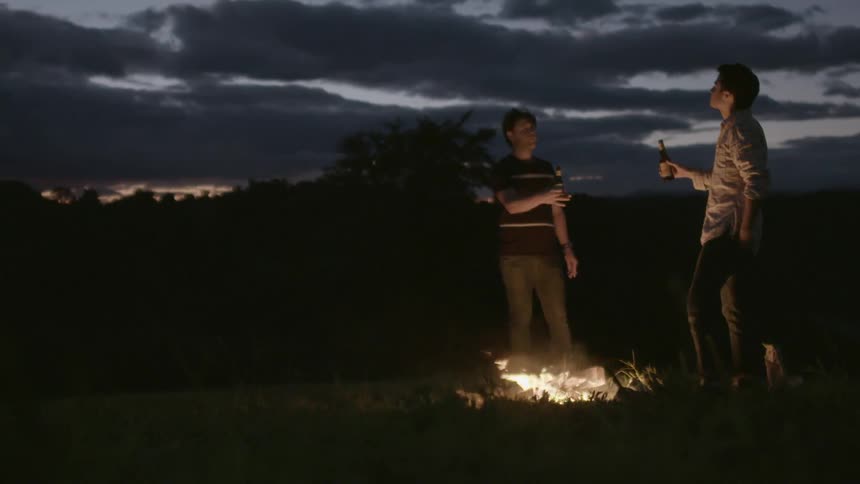 Two men stand and drink beers by a campfire at night