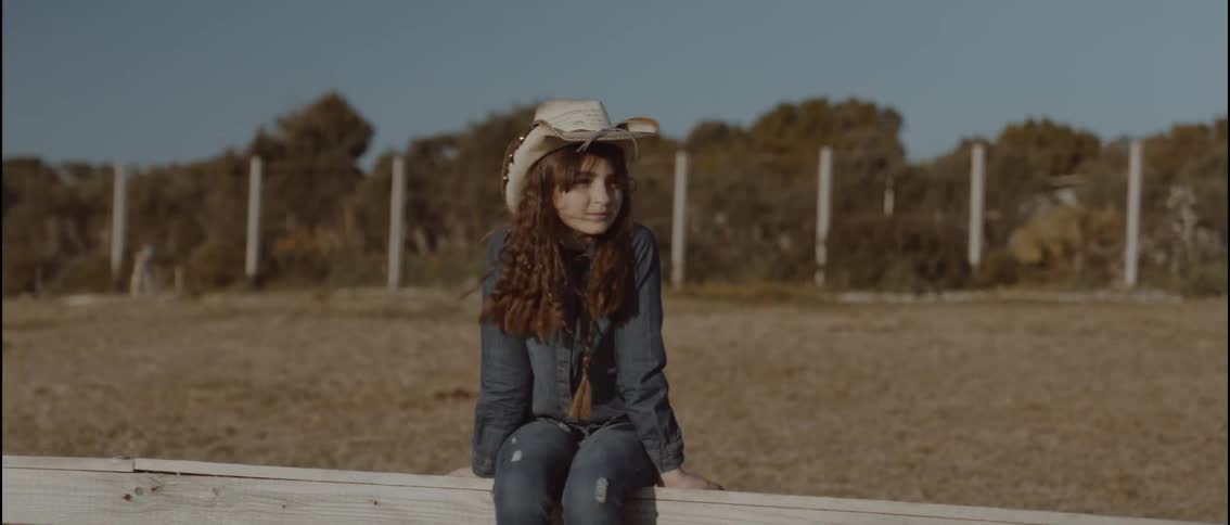 A young girl wearing a straw hat and denim clothing sits on a white wooden fence in a rural field during the day while a horse and rider pass by in the foreground
