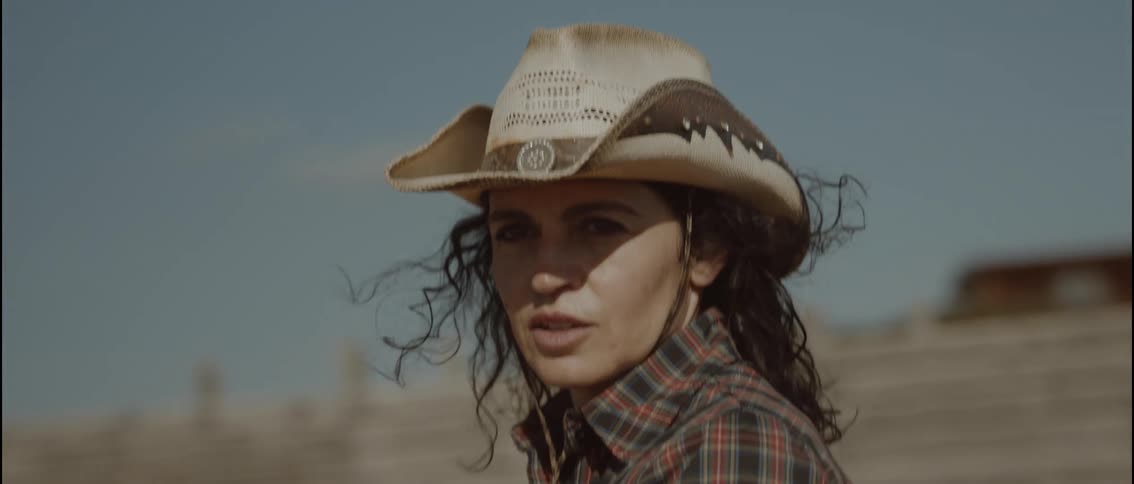 A woman wearing a straw cowgirl hat and a plaid shirt stands outside against a blurred ranch background
