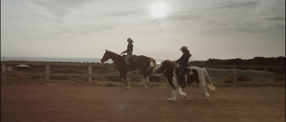 Two riders in cowboy hats gallop their horses along a dirt track overlooking the ocean at sunset the scene captures a brown horse and a spotted pony moving in parallel against a hazy golden horizon