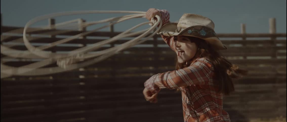 A young girl wearing a straw hat and a plaid shirt smiles and shouts while swinging a lasso in an outdoor arena with a wooden fence during the day