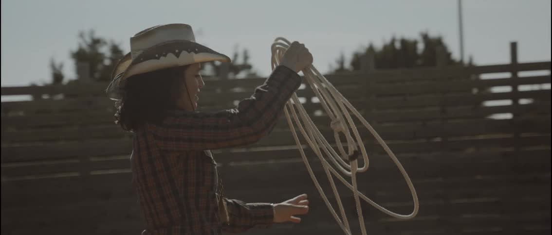 Young adult woman wearing a plaid shirt and cowboy hat roping next to a wooden fence with trees on the background outdoors during day time