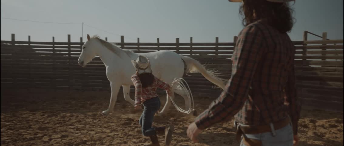In a dusty corral a young girl in a cowboy hat and plaid shirt runs alongside a white horse while swinging a lassoa woman stands in the foreground watching the girls progress as the horse trots along the wooden fence