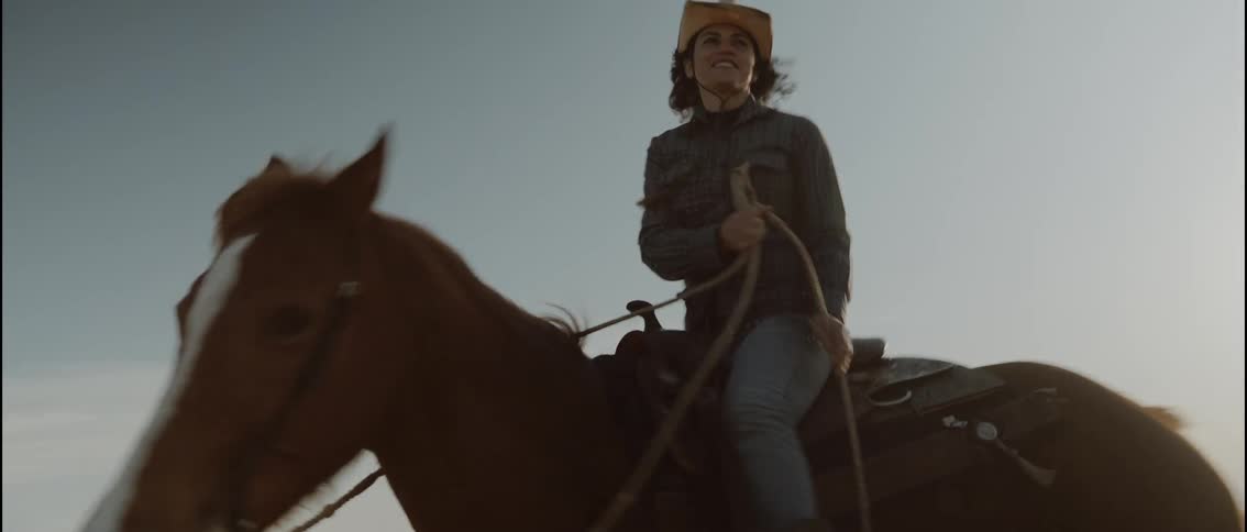 A woman wearing a cowboy hat and denim rides a brown horse across an open landscape she smiles and looks around while holding the reins as the horse moves at a steady pace under a clear sky