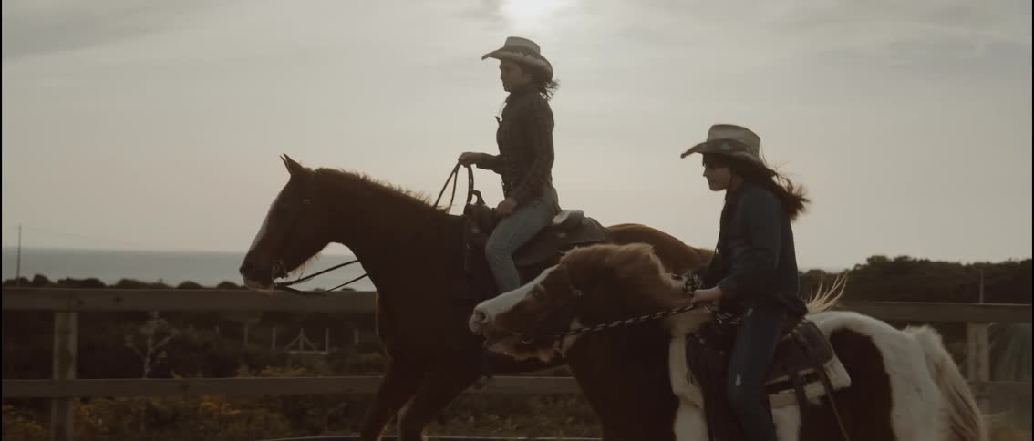 Young adult woman and teenager girl wearing plaid shirts and cowboy hats ridnig horses on a road next to a field close to the ocean outdoors during day time