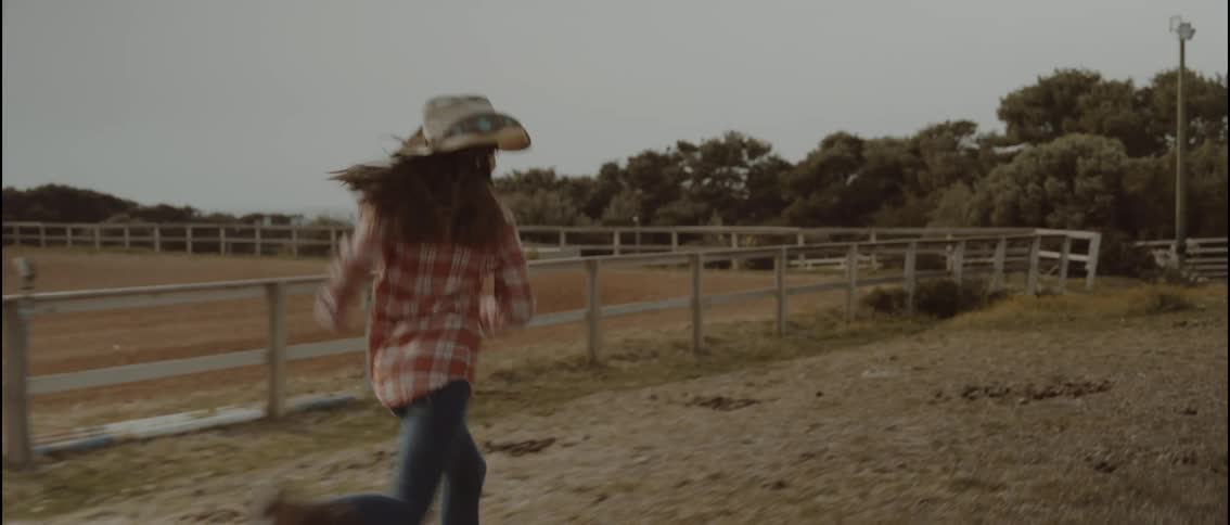 A young girl runs across a ranch at midday wearing a cowboy hat