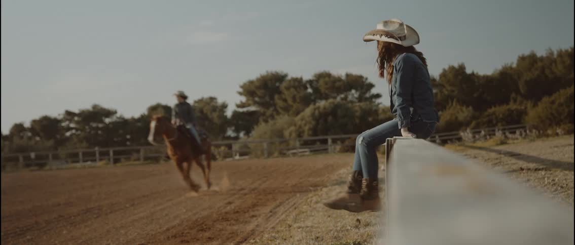 A girl wearing a cowboy hat and denim sits on a wooden fence while watching a woman riding a brown horse across a dirt arena under a clear sky