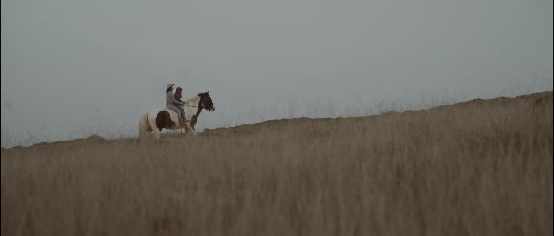 A woman and her daughter ride together on one big horse in a field of tall grass, without a saddle