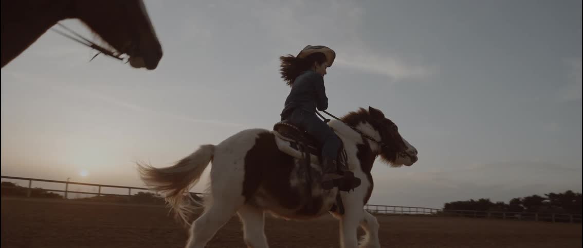 A young girl riding a pony while another person rides in the background in an arena at sunset