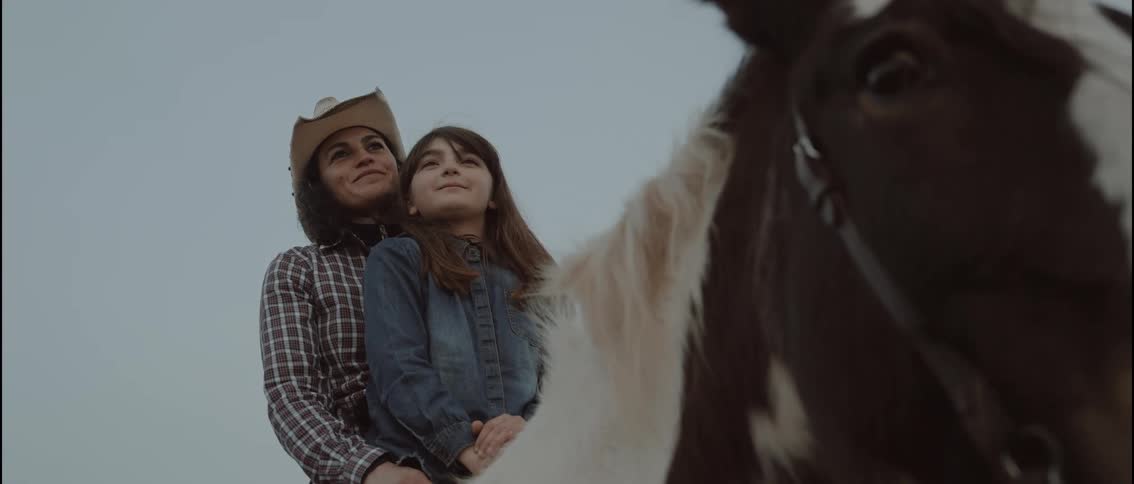 A woman and her daughter sit on a horse at sunset on a ranch with the woman wearing a hat