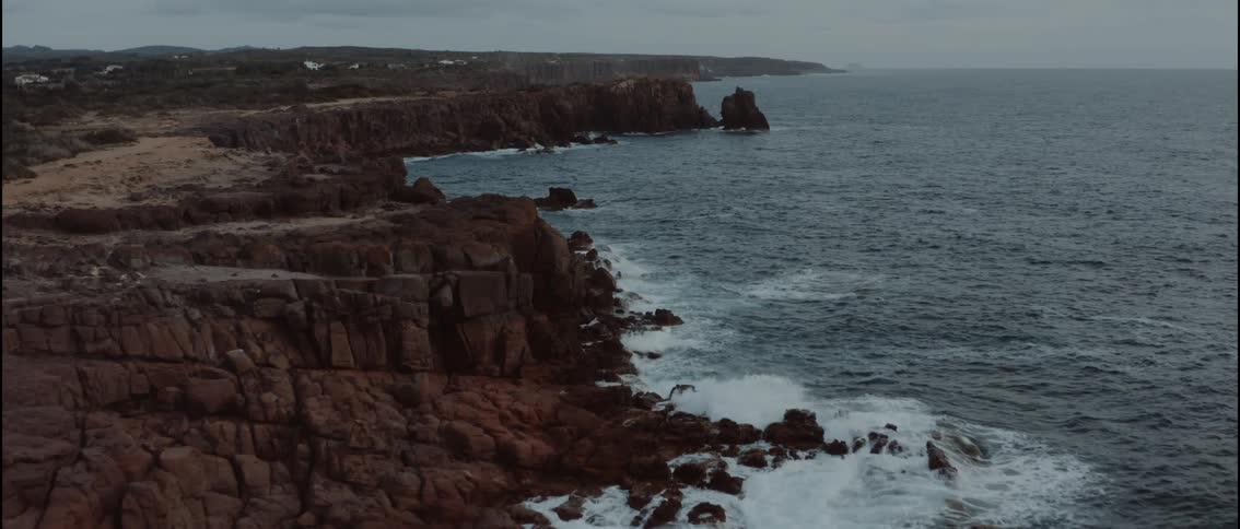 The video features an aerial shot of a rugged rocky coastline where dark cliffs meet the deep blue ocean white waves crash against the jagged rocks and outcrops under a vast overcast sky