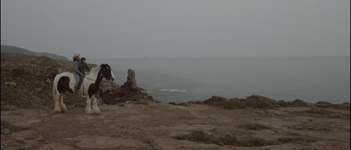Woman sits on a horse with her daughter along the coast as they look at the water