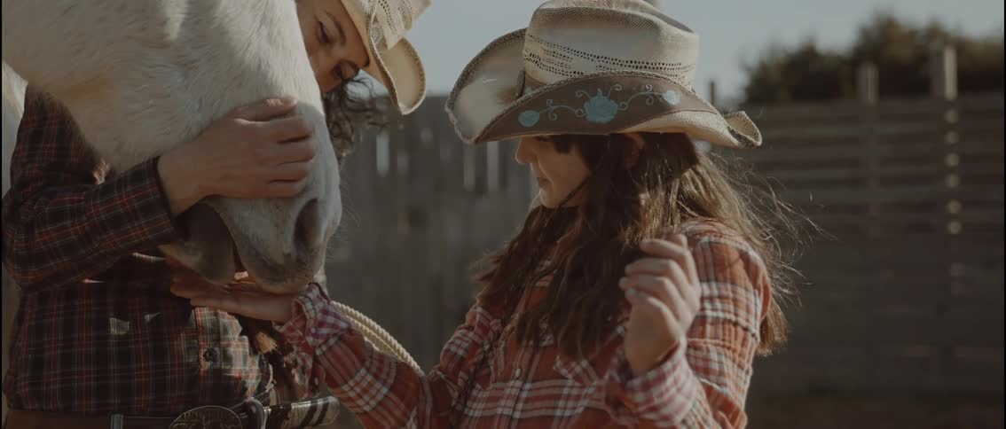 A woman and a young girl wearing cowboy hats are shown smiling and interacting affectionately with a white horsethe scene takes place outdoors near a wooden fence with the woman holding the horses head while the girl stands nearby laughing