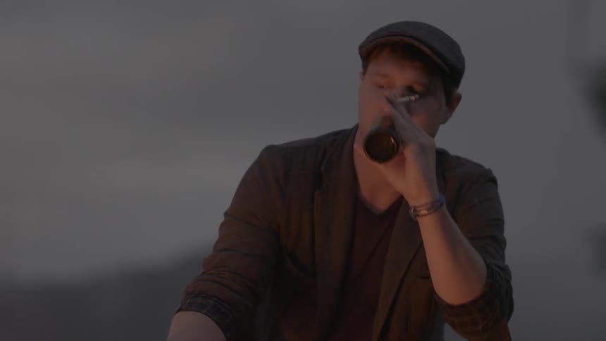 A man drinks beer and smokes a cigarette next to a campfire