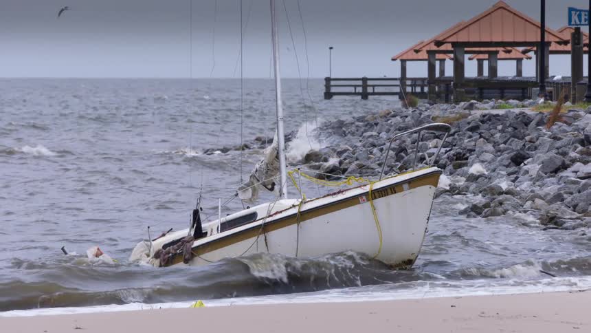 Sailboat stranded on the sand with strong waves hitting its side near a wooden pier