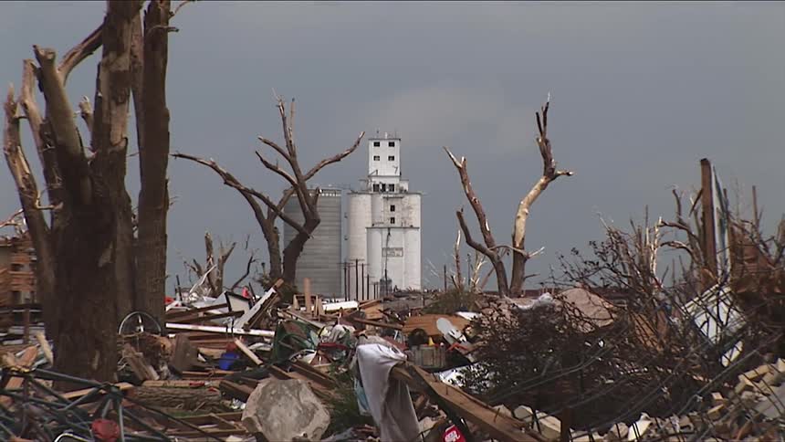 Man walking high on a tall building next to a silo among torn broken trees and destruction as a truck works to clear the debris