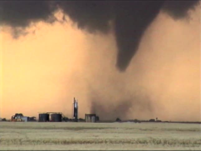 A large dark funnel cloud descends from a heavy storm shelf toward an open field the tornado kicks up a significant amount of dust and debris as it moves across the rural landscape near some farm structures