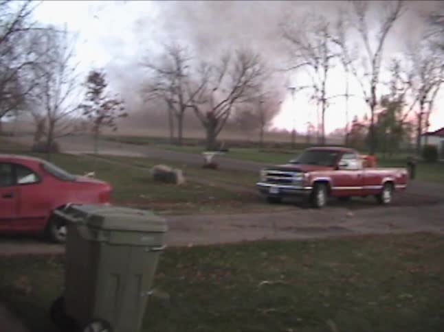 A large dark tornado lofts debris as it moves across an open field toward a residential areathe video captures the storms progression from behind a screen door showing cars parked in a driveway and bare trees swaying in the wind