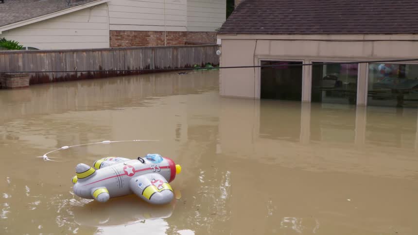 A backyard that has been completely overtaken by murky floodwaters reaching the windows of a nearby building in the center of the yard an inflatable toy airplane and a blue floatie drift slowly across the submerged space