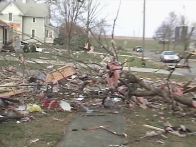 People walking outside looking at tornado destruction in a neighborhood with some houses destroyed and others untouched with trees broken and torn