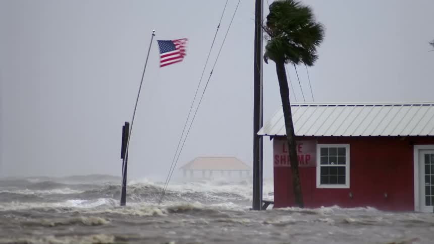 On a windy cloudy day in a suburban area floodwaters rise periodically above ground level as strong winds with a building a tree and a flagpole in view the flag whipping in the wind