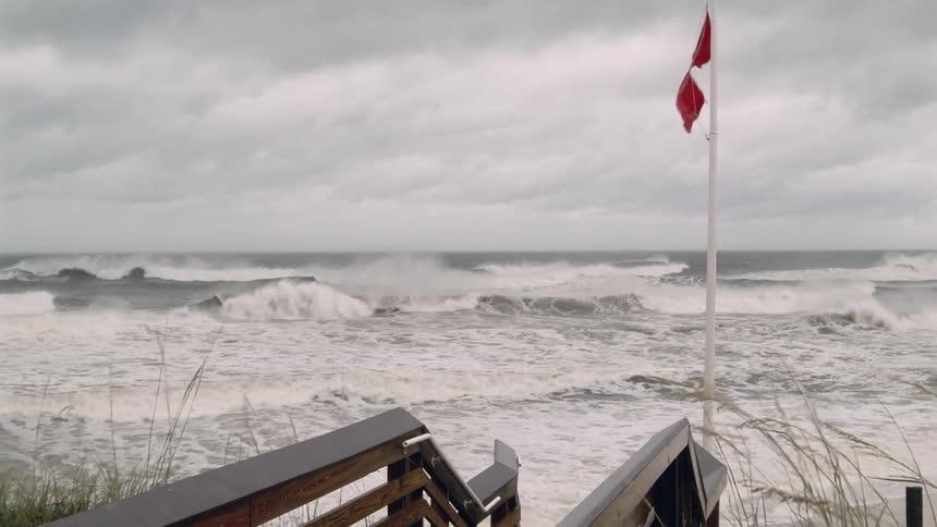 Two red flags whip violently in the wind atop a tall pole signaling extremely dangerous conditions as rough whitecapped waves crash onto the shoreovercast grey clouds hang low over the churning atlantic while a wooden boardwalk path leads the eye t