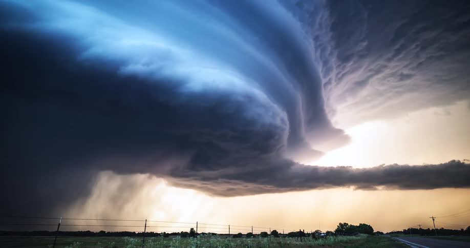 A massive supercell storm rotates over a rural landscape displaying dramatic shelf cloud layers and heavy precipitationthe sky transitions from a deep electric blue to a fiery orange glow as the sun sets behind the advancing storm front