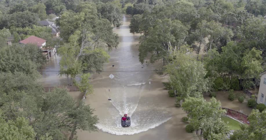 An aerial shot shows a small boat navigating through a suburban street completely submerged in deep murky floodwaterslush green trees and the rooftops of houses line the flooded roadway highlighting the extent of the natural disaster