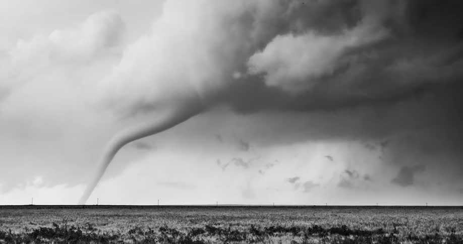 Elongated tornado extending beneath dense clouds over flat field