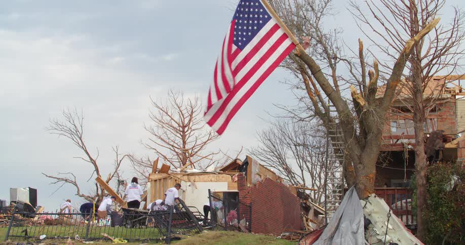 A large american flag waves from the broken limb of a tree in the foreground overlooking a group of people in white shirts sifting through the massive piles of timber and debris from a flattened home