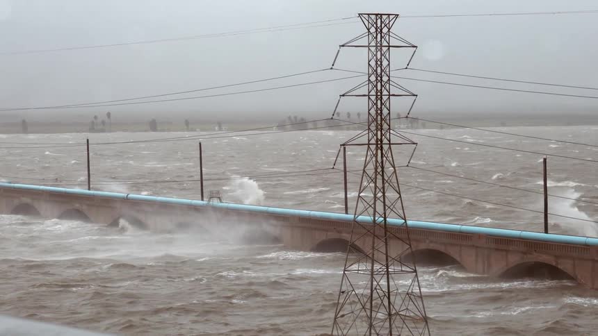 Violent storm surges and whitecapped waves crash against a stone arched bridge during a period of heavy raina tall power line tower stands in the foreground as wind and water spray lash across the gray turbulent landscape