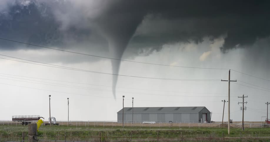 Narrow tornado forming near a barn in a rural area with utility poles and power lines