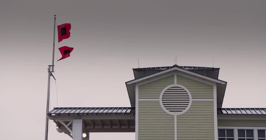 The video shows a pale green building with a prominent circular vent on its facade situated under a dark and overcast sky to the left two red flags with black squares in the center flap in the wind on a tall flagpole signaling a hurricane warning