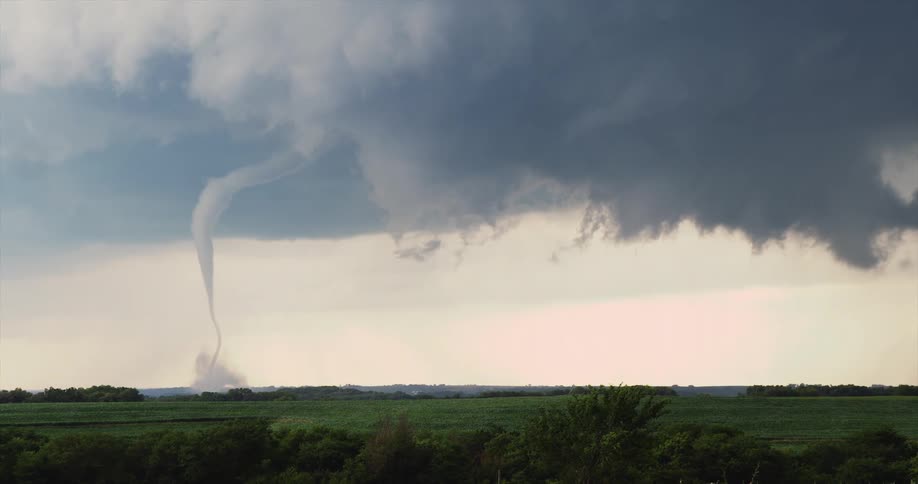 On a cloudy day a tornado forms in the distance over a grassland with dark clouds covering the sky as lightning briefly strikes