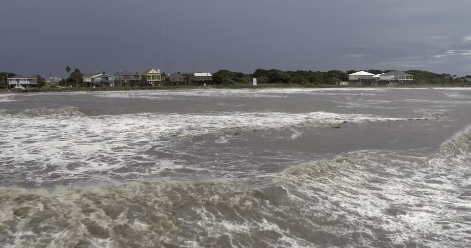 Houses on stilts in a coastal community as rough water waves roll in to shore under a dark sky