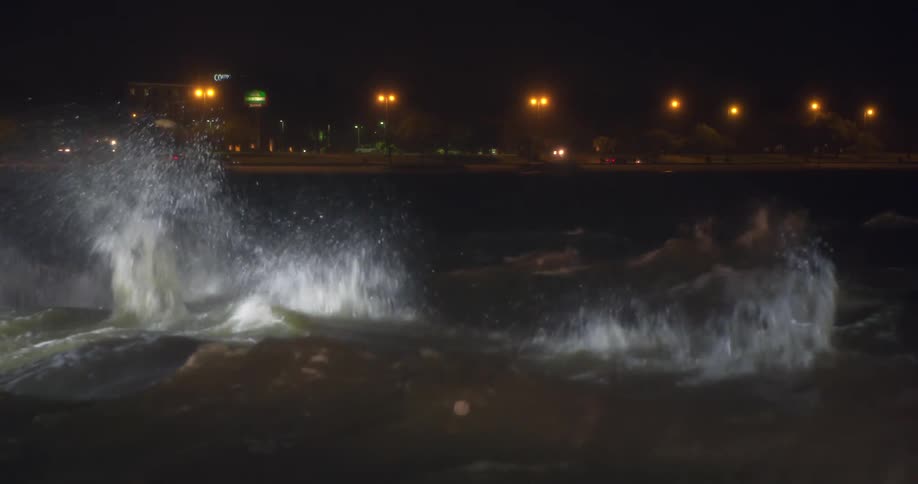 Ocean waves crash onto the shoreline at night with a road along the coast illuminated by streetlights as cars pass by