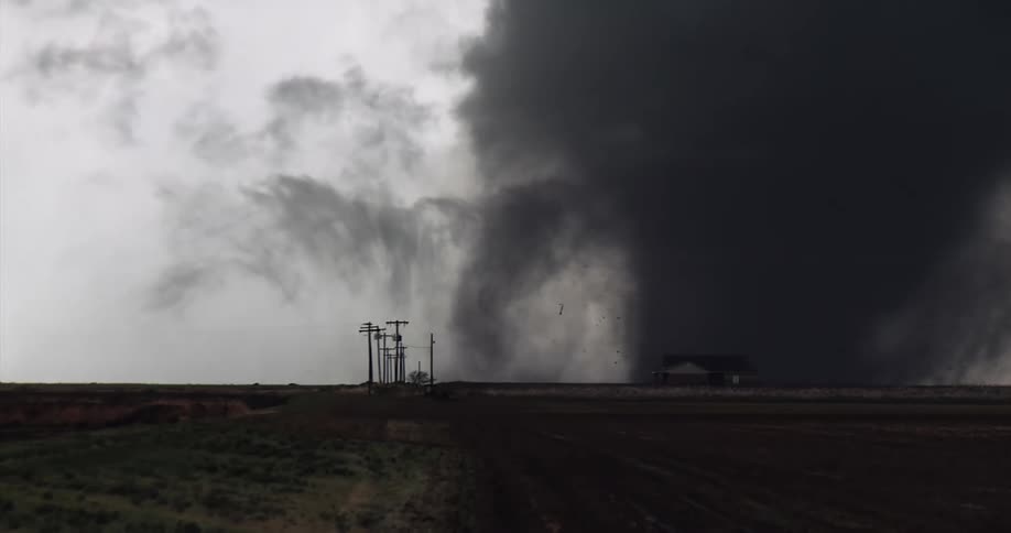 A massive dark tornado looms over a flat rural landscape its violent rotation churning up debris near a small farmhouse and power linesthe heavy sootcolored clouds of the funnel contrast sharply with the pale overcast sky to the left creating a s