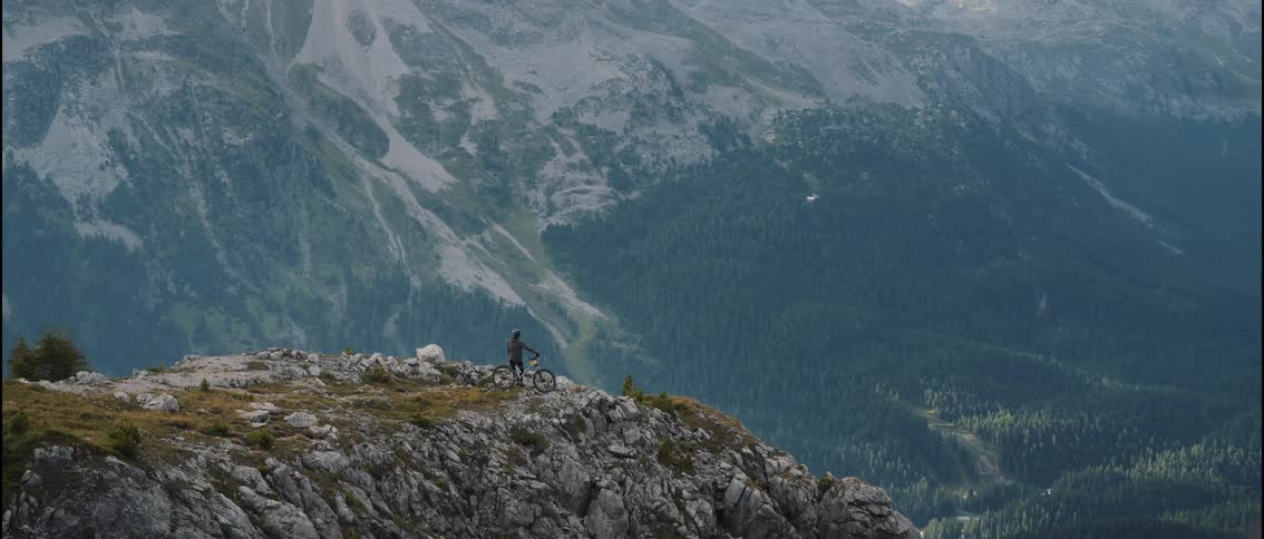 A lone hiker stands still on a small mountain peak while much larger mountains rise behind them creating a layered view of rugged terrain and open sky that emphasizes the scale of the surrounding wilderness