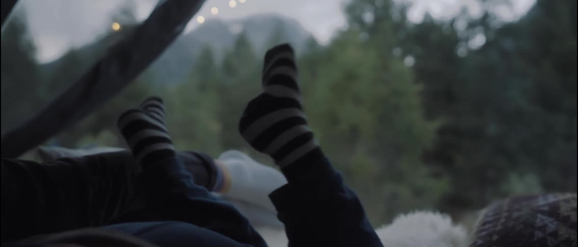 A person wearing striped socks gently sways their feet inside a tent while another figure rests nearby with soft string lights glowing against a blurred forest and mountain backdrop beyond the open tent