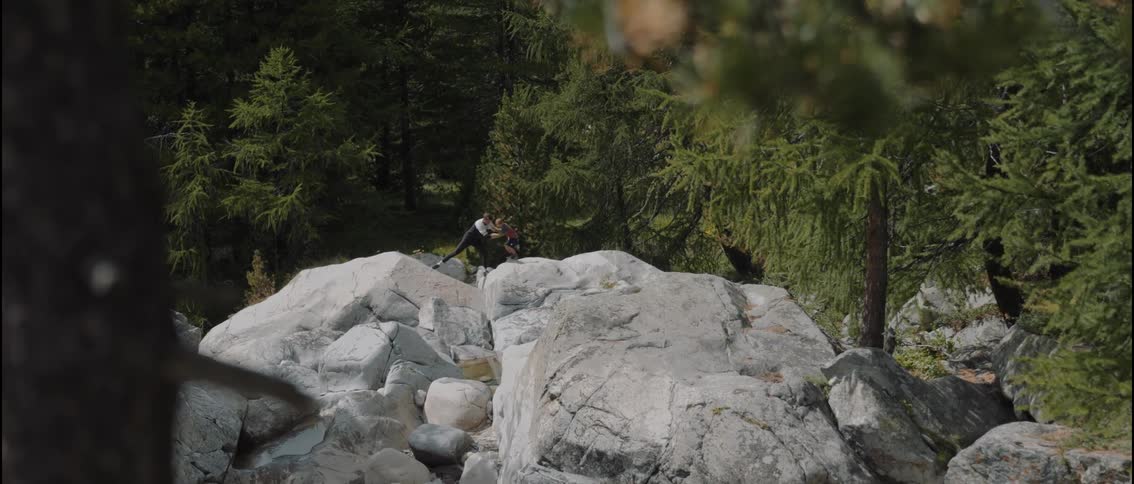 A father and son walk and climb over rocks in the forest as the father lifts the boy from one rocky surface to another