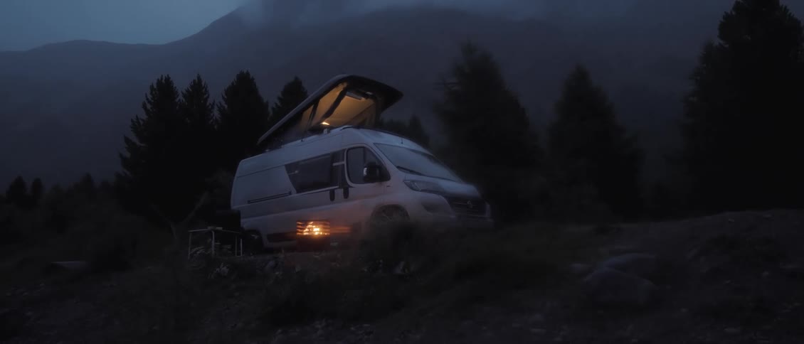A white camper van with string lights on its pop up roof sits in a mountain forest at night as the lights turn off