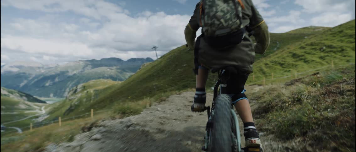 A child wearing a helmet and backpack rides a mountain bike down a winding dirt trail through rolling green hills beneath a partly cloudy sky