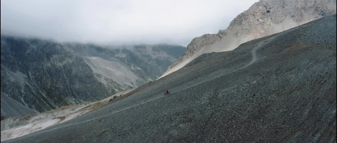 A lone cyclist rides across a vast rocky mountain slope beneath lowhanging clouds that cling to the rugged peaks
