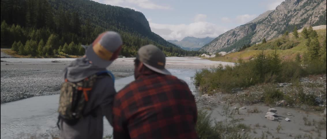 A man wearing a plaid shirt and a young boy wearing a backpack sit together by a wide mountain river and point toward the valley and evergreen forests under a bright sky