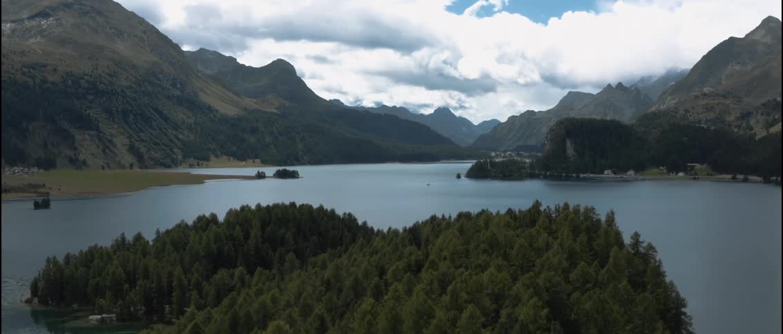 A vast blue lake surrounded by dense evergreen forests and steep mountain peaks under a cloudy sky